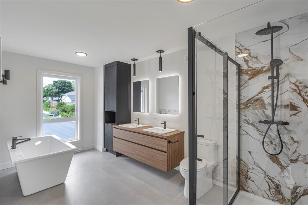 Salle de bain d'une maison neuve : bain autoportant, vanité double en bois, miroirs rétroéclairés et douche en céramique effet marbre